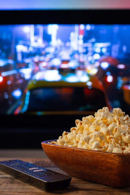 A wooden bowl of popcorn and remote control in the background the TV works. Evening cozy watching a movie or TV series at home.